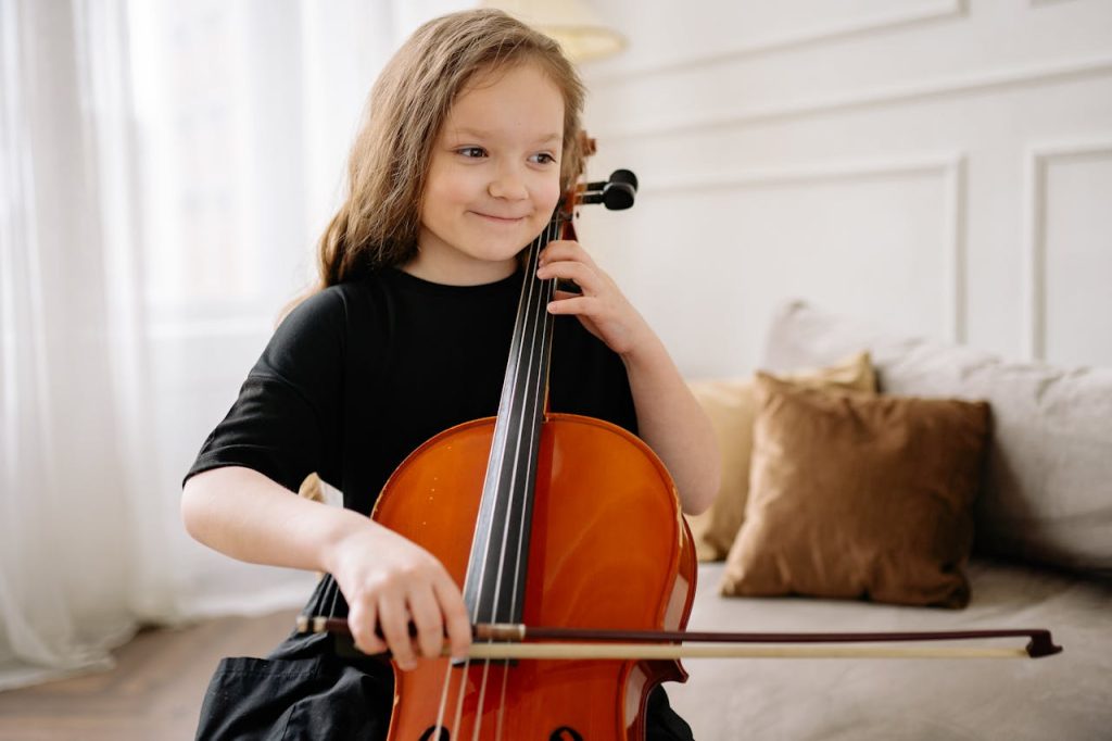 A cute girl in a black dress plays the cello indoors, smiling happily.